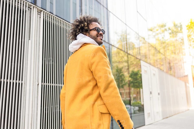 A stylish afro man in a vibrant yellow coat and sunglasses walks confidently past a modern glass building, embracing a casual yet fashionable look in an urban setting
