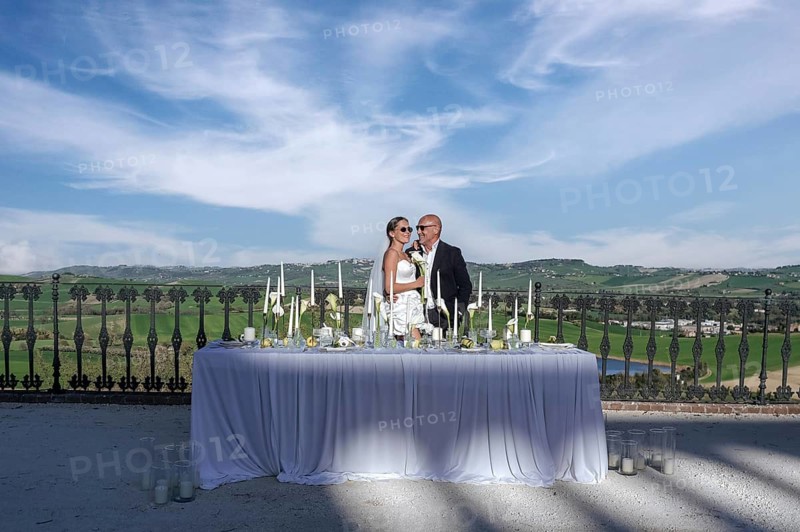 A romantic wedding scene in an ancient villa in Treia, Italy, with a breathtaking view over the rolling hills of Macerata. The bride and groom stand beside an elegant setup