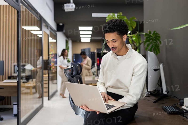 A professional office environment with a man using a laptop while sitting on a desk, and colleagues collaborating in the background, surrounded by modern decor and technology