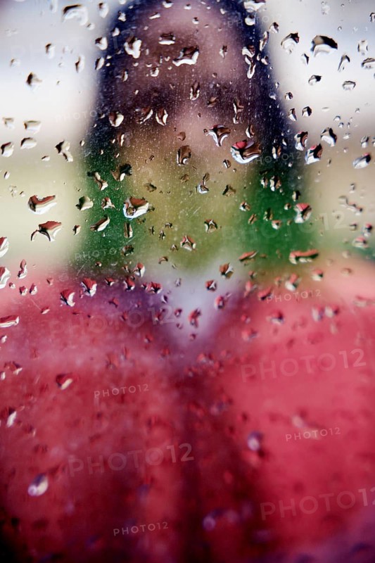 A blurred silhouette of a person visible behind a rain covered glass window. Droplets create an abstract pattern, contrasting with the vibrant colors in the background