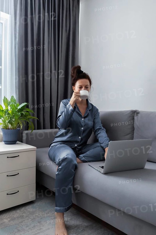 A woman sits comfortably on a gray sofa, wearing blue pajamas. She is enjoying a cup of coffee, her laptop open beside her. The cozy scene is perfect for remote work or relaxation themes