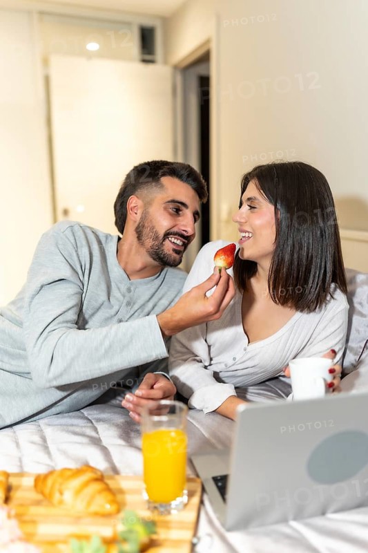 Loving couple sharing a strawberry while having breakfast in bed with a laptop, enjoying a romantic and connected morning