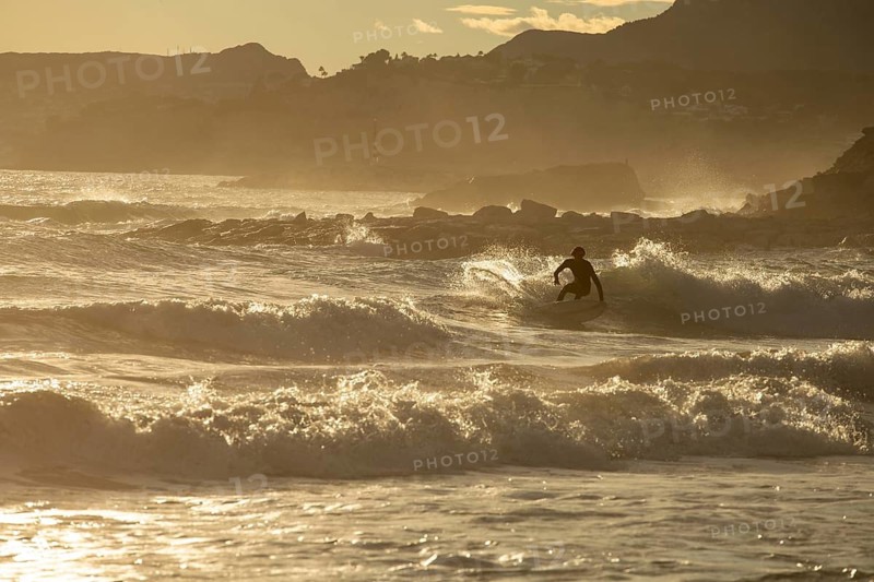 A lone surfer rides the waves against the stunning backdrop of the Costa Blanca, with the golden hues of a Mediterranean sunset casting a serene glow on the sea and landscape