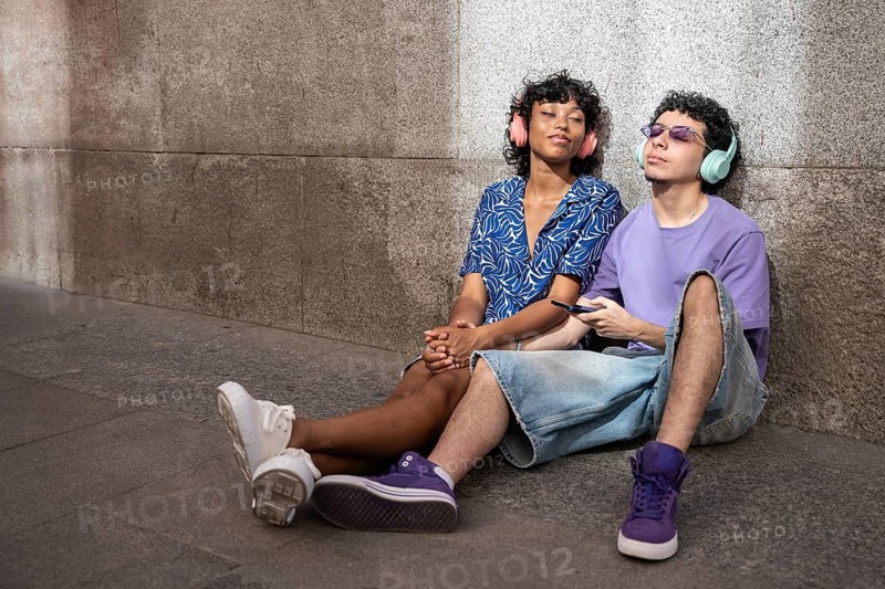 A joyful multiethnic LGBTQ+ couple listens to music with headphones while sitting against a concrete wall in a sunlit urban area, enjoying each other's company