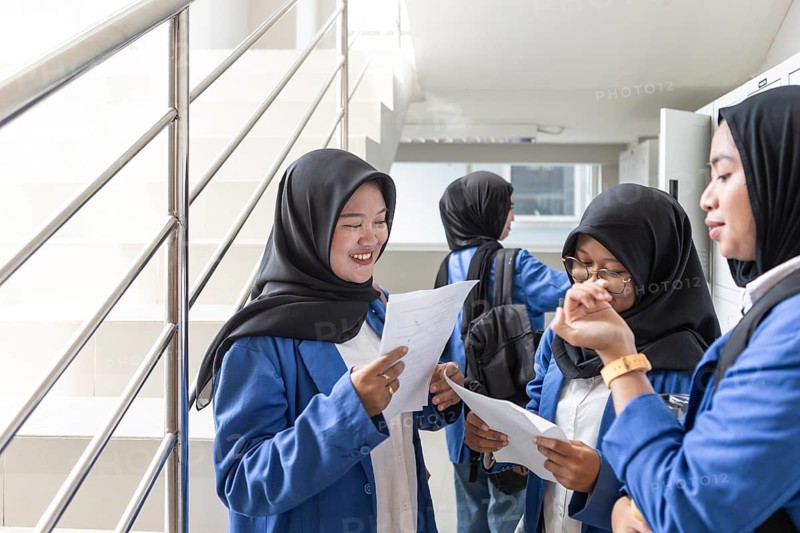 Asian Muslim students in hijabs engage in a lively discussion, holding papers, as they prepare for school activities. They are wearing blue uniforms in a bright hallway