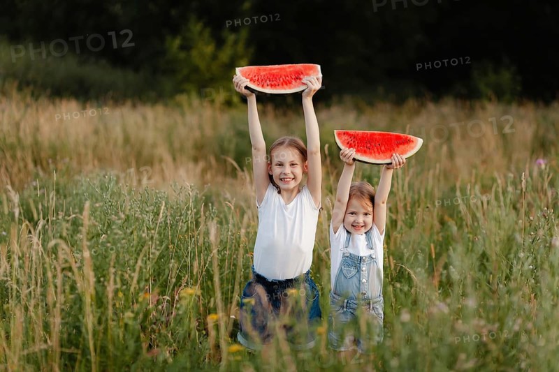 Two cheerful children joyfully raise watermelon slices in a lush field, capturing the essence of summer fun, freshness, and innocence amidst the bounty of nature and sunshine