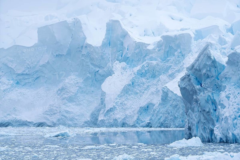 Glacier eruption edge with shimmering blue ice formations, in front of iceberg and floating ice floes on calm water surface, Andvord Bay, Antarctic Peninsula, Antarctica