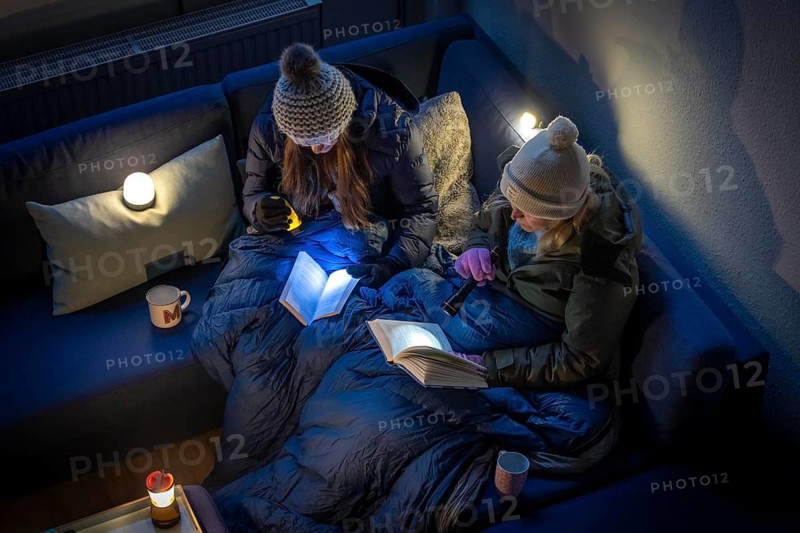 Symbolic picture blackout, extensive, prolonged power failure, in private surroundings, without electricity, heating, water, family on the sofa with warm winter clothes, sleeping bag, light from battery lights, battery lights, flashlights
