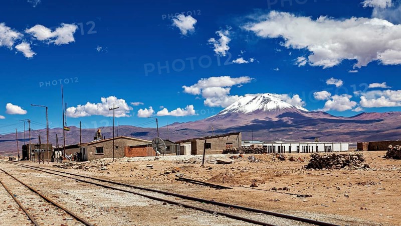 Houses next to train tracks with an impressive volcanic background under clear skies, the town of Ollagüe in the Chilean Atacama Desert with the Ollagüe volcano of the same name in the background