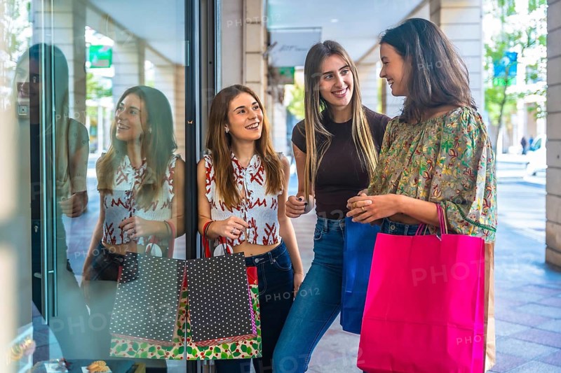 Three smiling young women chat outside a boutique storefront, holding colorful shopping bags, laughing and enjoying a sunny downtown shopping outing together