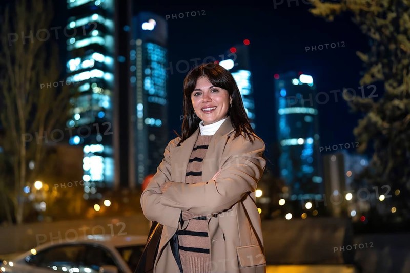 Young woman standing confidently with arms crossed, smiling at the camera outdoors at night, with illuminated city skyscrapers and bokeh lights in the urban background
