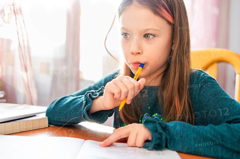A young girl in a blue sweater sits at a desk, focusing intently on her homework. She holds a pen to her lips, appearing deep in thought, surrounded by books