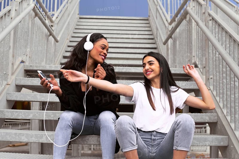 Two friends sit on outdoor stairs, sharing earphones and music from a smartphone. They appear cheerful and relaxed, highlighting friendship and modern technology