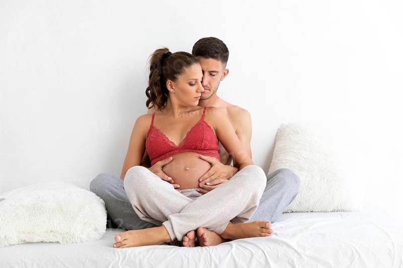 An expectant couple embraces lovingly while seated on a bed, highlighting the warmth and anticipation of parenthood The background is a simple, soft white setting