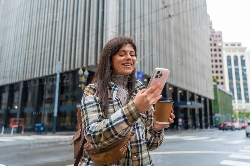 Young woman standing on a city street, holding a coffee cup and smiling while actively using her smartphone, showcasing urban digital connectivity and a refreshing break in san francisco