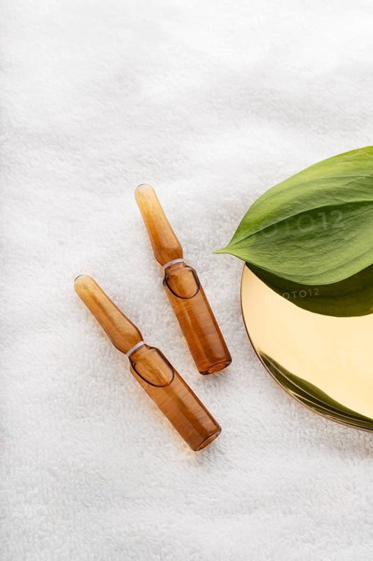 Two amber glass cosmetic ampoules filled with anti-aging serum, accompanied by a fresh green leaf on a soft, textured white surface