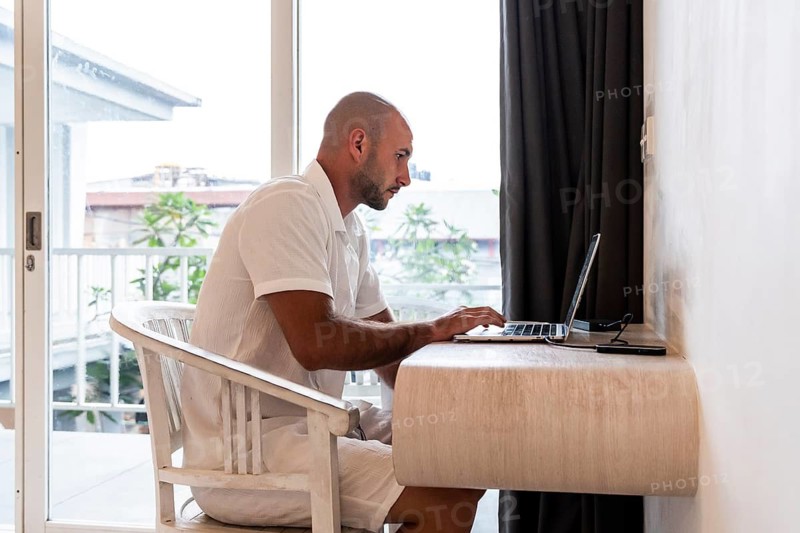A man in a white shirt sits at a desk in a hotel room, focusing on his laptop. A sliding glass door opens to a balcony with a view, blending work and leisure