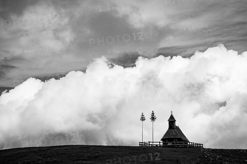 A traditional small wooden chapel with a cross sits on a green grassy ridge under a bright blue sky with large white fluffy clouds in Slovenia