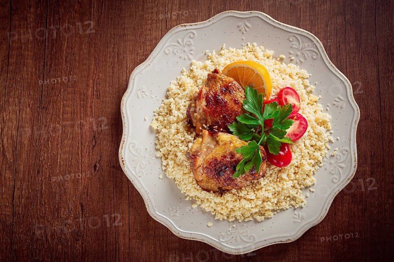 Couscous is served on a plate with pieces of chicken on top. Lemon slices and parsley decorate the dish