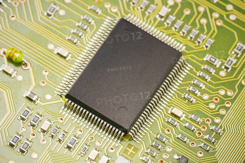 A detailed view of a microchip positioned on a green circuit board. The intricate paths and components highlight the complexity of modern electronic devices