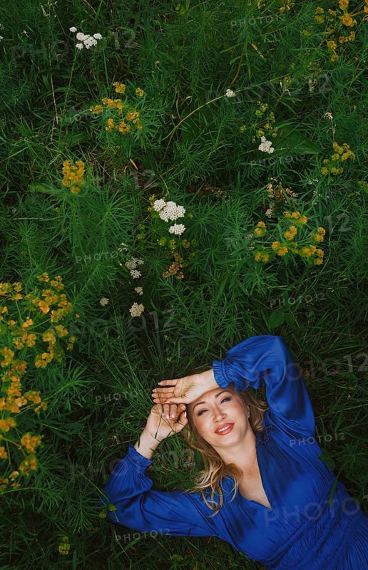 A cheerful blonde woman lies joyfully in a grassy field, surrounded by vibrant green foliage and wildflowers. She wears a blue dress, expressing freedom and serenity
