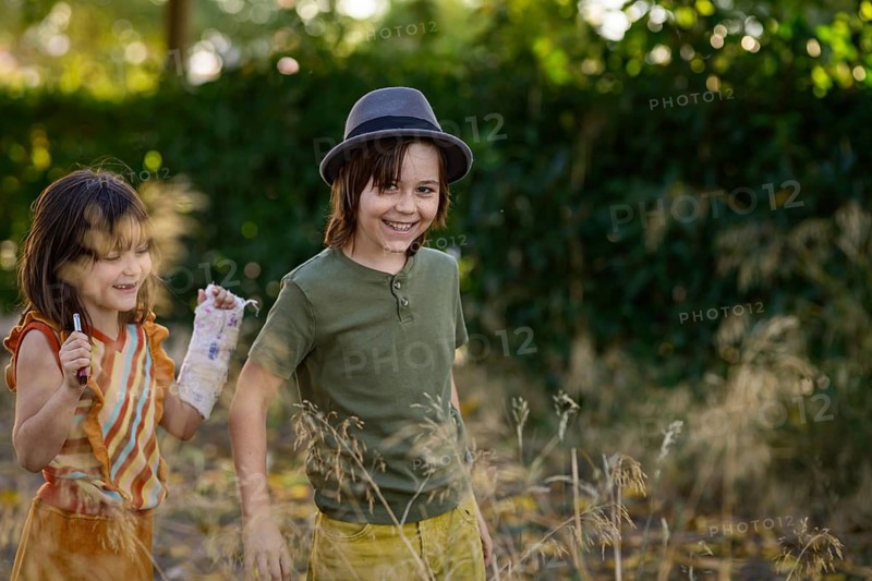 Two children happily play in a sunlit garden, surrounded by tall grass and greenery. The boy wears a gray hat, while the girl holds a decorated cast and pen