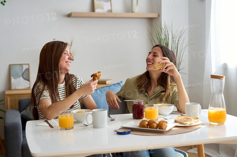 A lesbian couple shares a joyful breakfast at home, surrounded by light and warmth They enjoy croissants and orange juice while chatting and laughing together