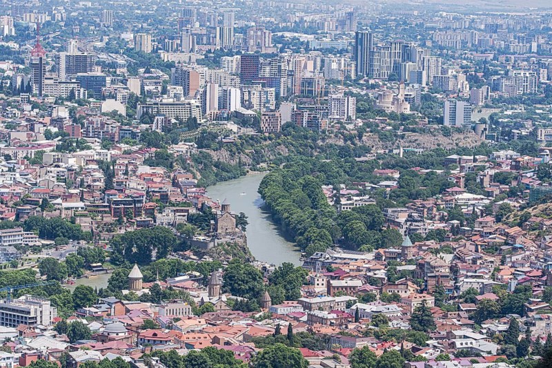 Aerial view of Tbilisi showcasing a blend of historic and modern architecture. The Kura River winds through the city, bordered by lush greenery and iconic landmarks