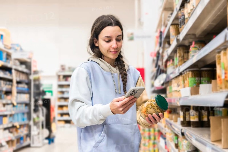 A woman in a supermarket is intently looking at her smartphone while holding a jar of food. She is browsing or reading something on the phone, possibly comparing prices or checking a shopping list