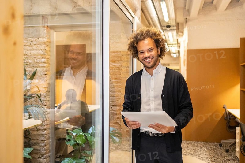 A smiling man with a tablet stands in a modern coworking space, showcasing a bright, collaborative work environment. The setting features natural light and contemporary decor