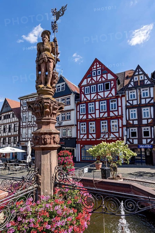 Market fountain man with market flag, knight, sandstone sculpture, fountain figure at the Kump, medieval market fountain with geraniums, decor, narrow high half-timbered houses on the market square, pedestrian zone, historic old town, Butzbach, Wetterau, Hesse, Germany