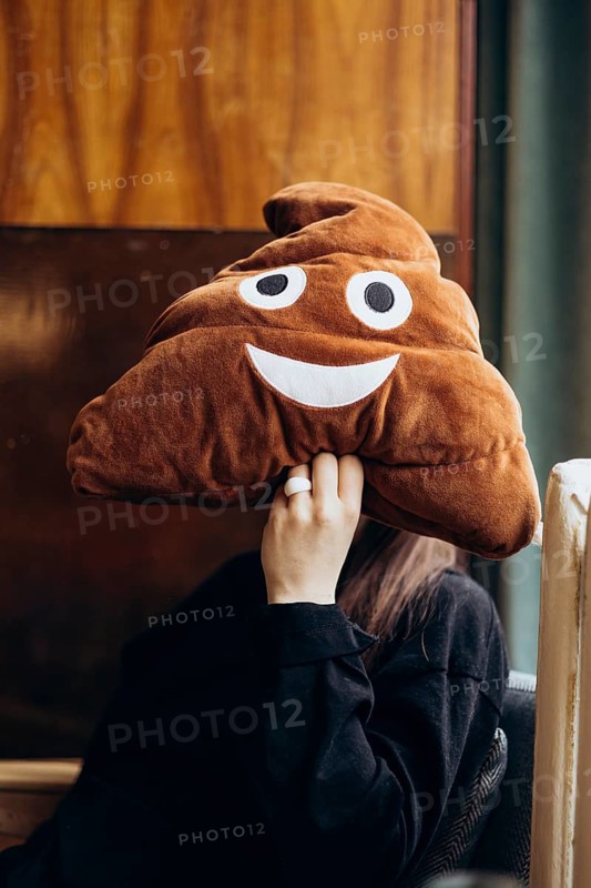 Cropped unrecognizable young woman holding a large plush poop emoji toy over her face in a cafe, adding humor and anonymity to the scene