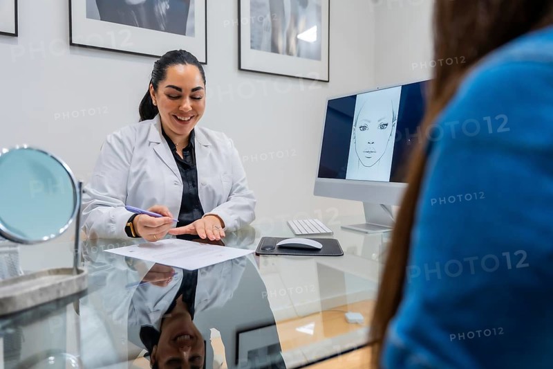 Female doctor in a white coat consulting a young woman about potential aesthetic beauty procedures or plastic surgery, showing face sketch designs on a computer monitor