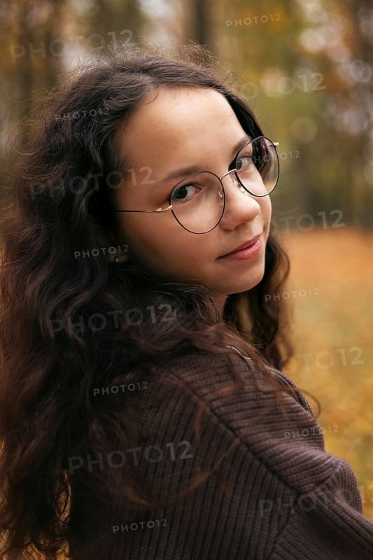 A teenage girl with long curly brown hair and glasses looks over her shoulder in an autumn forest. She wears a cozy brown sweater, blending with the warm fall colors around her