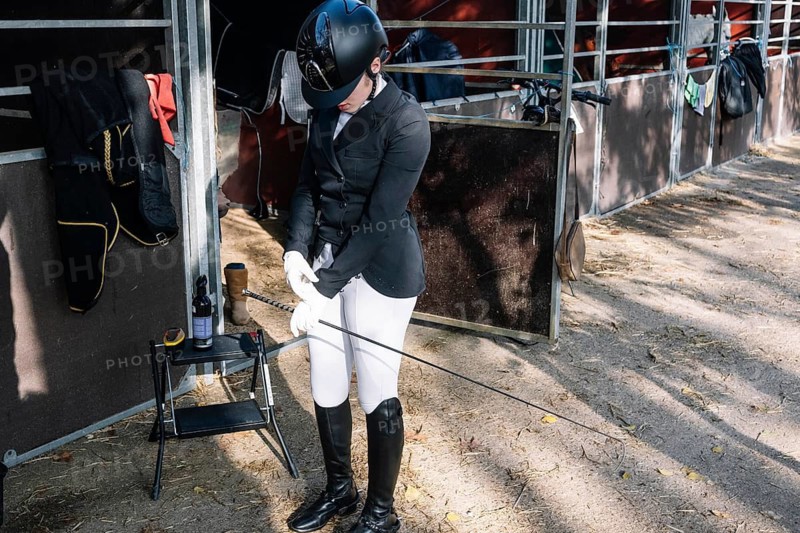 A teenage female equestrian dressed in classical dressage attire adjusts his gloves. She is looking down, captured beside stable gear, showcasing a typical pre-competition scene