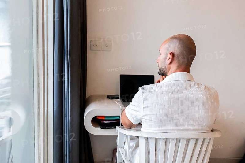 A man seated at a desk in a hotel room, working on a laptop. The bright interior and modern furnishings create a productive environment for remote work