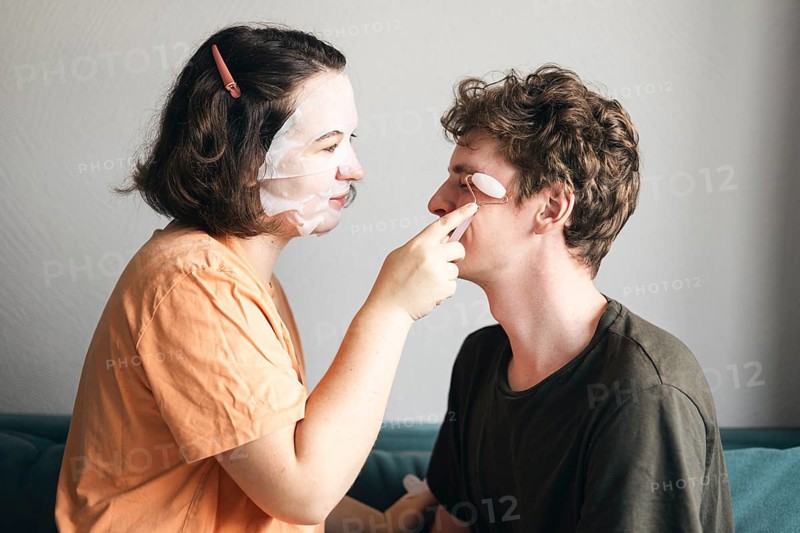 A young man and woman enjoy a playful skincare routine together in their living room. Both are smiling gently, she is covered in face masks, using a pink quartz roller on his face, underlining their cozy, intimate moment at home