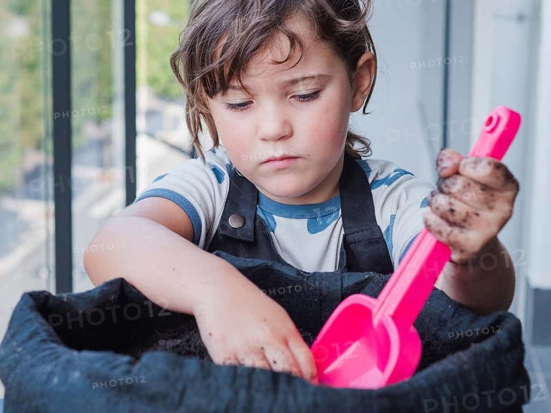 A focused young girl is seen planting a sunflower seed in a dark planter with a pink shovel, her hands soiled with dirt as she engages in gardening