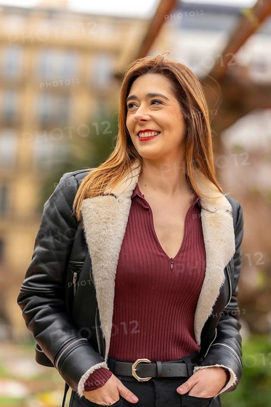 Young female journalist smiling and looking away while posing with hands in pockets in a blurred urban city setting