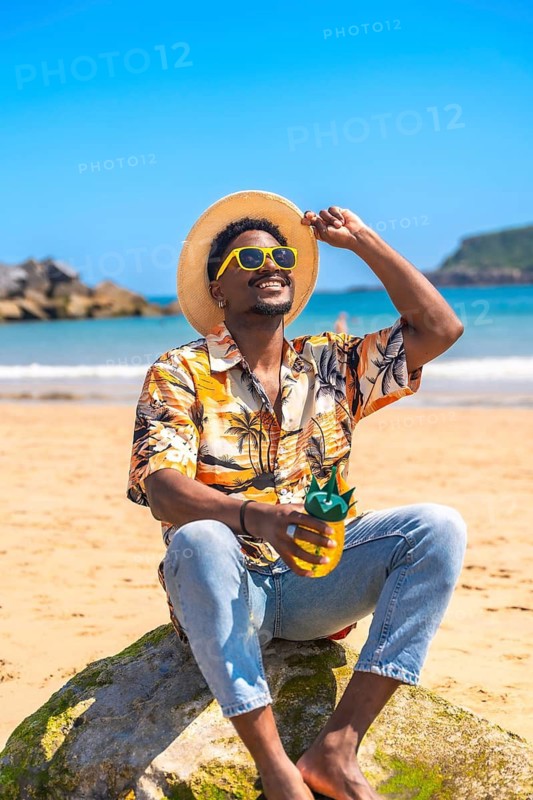 Young man wearing sunglasses and straw hat holding a pineapple shaped cocktail while relaxing on a rock at a sunny beach
