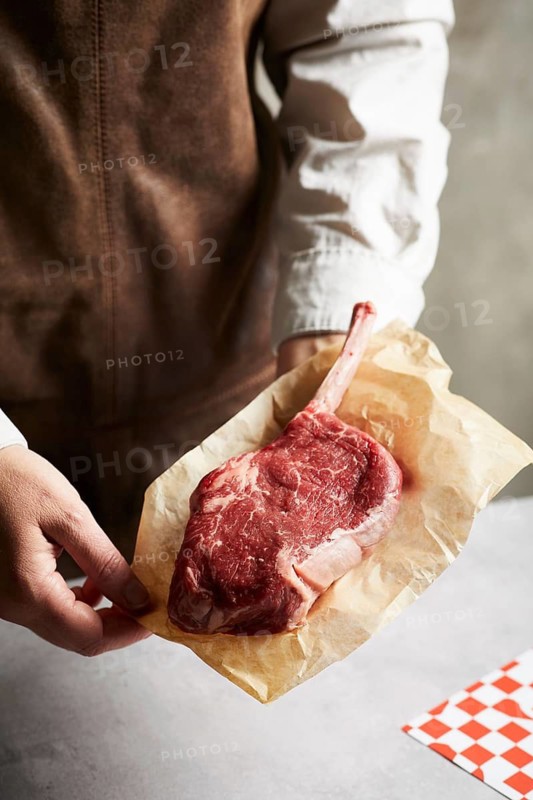 Close-up of a butcher or chef holding a raw tomahawk steak wrapped in brown butcher paper. The thick, bone-in cut is ideal for grilling or gourmet meat preparation