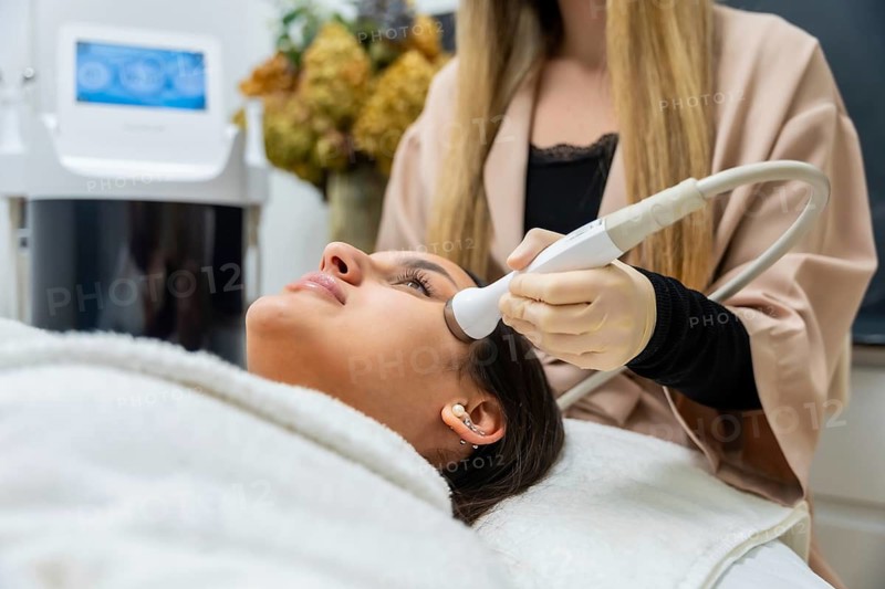 Woman lying on a bed receiving an aesthetic facial treatment from a professional using a modern device, focusing on skincare, beauty, and wellness in a clinic environment