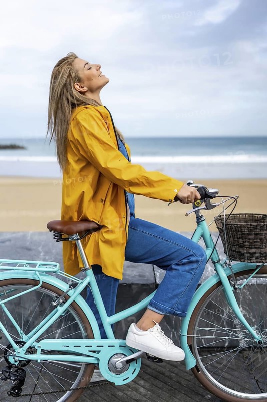 Young woman wearing yellow raincoat enjoying fresh air while riding bicycle on a cloudy day by the sea