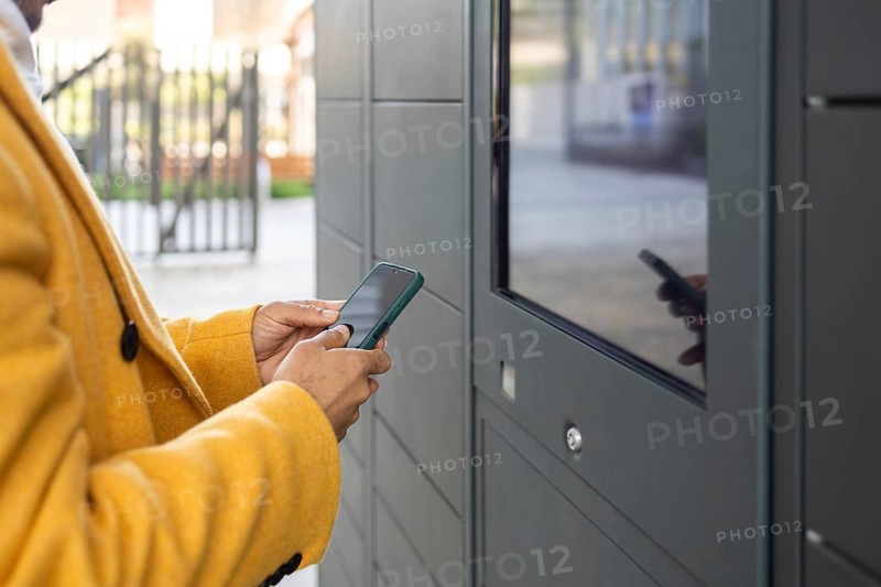 A businessman in a yellow coat uses a smartphone at an outdoor digital kiosk. The setting is urban and modern, suggesting a focus on convenience and technology in daily life
