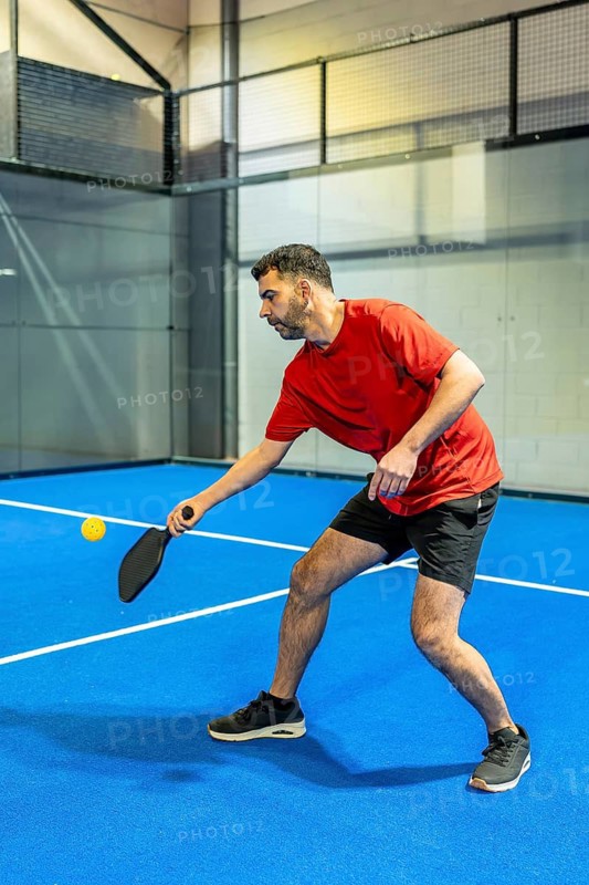 Male athlete in a red shirt striking a ball with a paddle on a vibrant blue pickleball court, mid action during a focused indoor game showcasing speed, skill and fitness