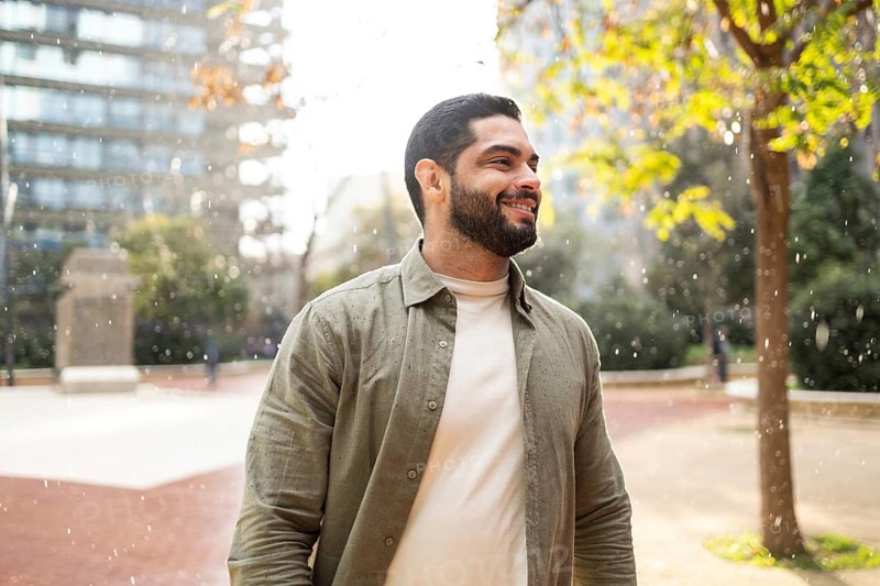 A man with a beard smiles while enjoying a light rain in a park of the city. He wears a light jacket, surrounded by trees and modern buildings, capturing a moment of joy