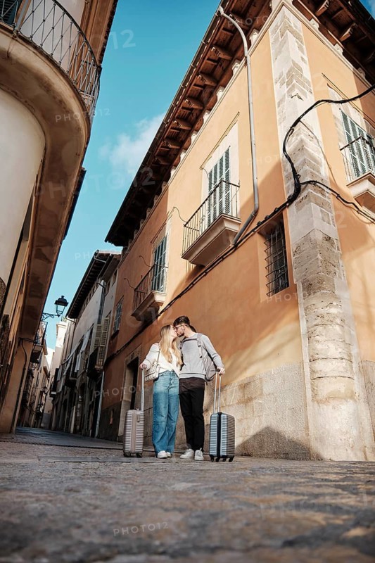 A young couple strolls with luggage in a charming Majorcan street, searching for their dream vacation home. Sunlight casts soft shadows on the warm-colored buildings