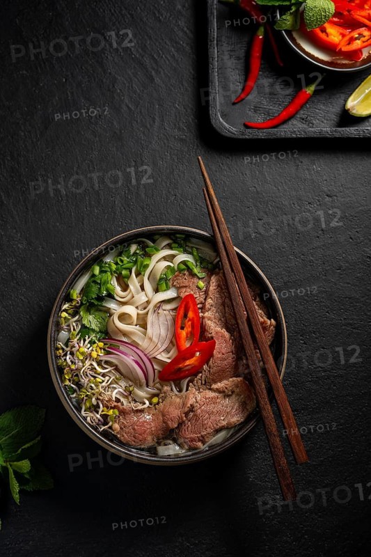 Top view of a delicious bowl of Vietnamese Beef Pho Noodle Soup, featuring tender slices of beef, flat rice noodles, fresh herbs like cilantro and basil, and vibrant garnishes of lime, red chili, and sprouts presented on a textured dark background