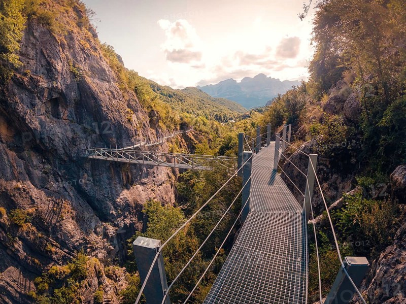 A metal bridge spans a scenic mountain gorge in the Pyrenees, surrounded by lush greenery and rocky cliffs under a vibrant sky, creating a picturesque and dramatic natural setting in summer