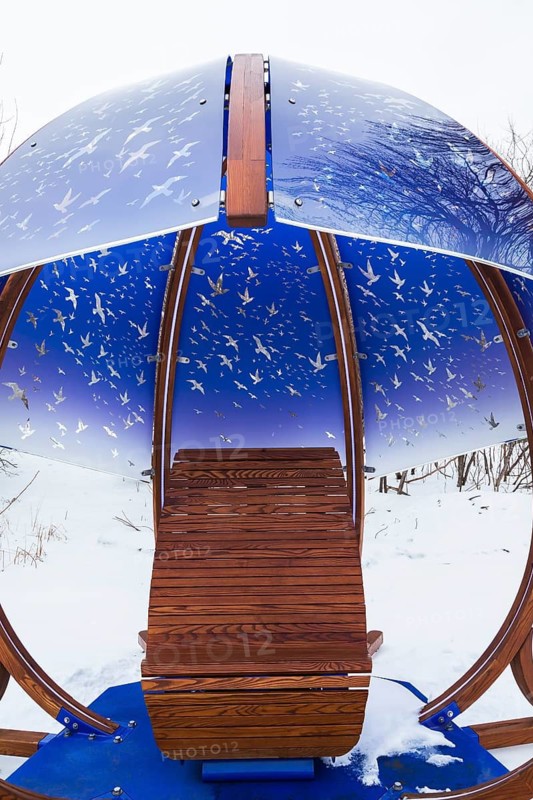 Interactive cocoon rocking chair made of curved wood and backlit blue tinted plexiglass panels canopy decorated with flock of birds in flight theme motifs created by Olivier Landreville and installed in winter like snow in early spring, Horizon Art Installation, Ile des Moulins, Old Terrebonne, Quebec, Canada